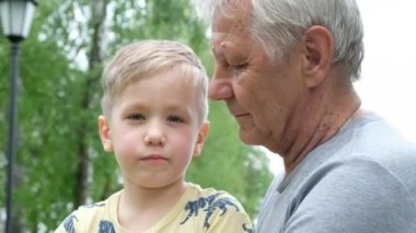 Küçük çocuk büyükbabasına sarılıyor ve onunla konuşuyor. Yetişkin bir adam, dışarıda parkta oturuyor.