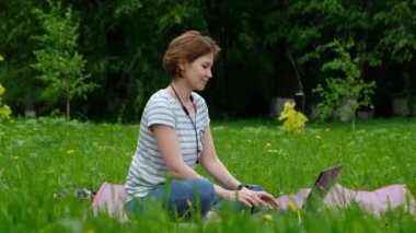 Genç bir kadın parkta oturuyor ve dışarıda bilgisayarla uğraşıyor.