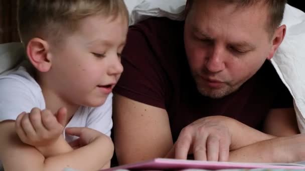 Far pappa och hans son ligger på en säng och läser en bok. Familjebegreppet — Stockvideo