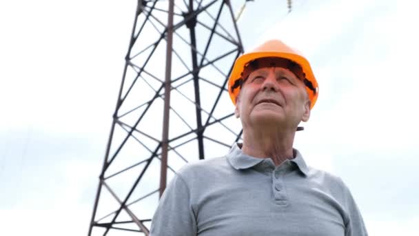 Senior man Chief Engineer in the Hard Hat looking at camera, by Transmission Tower, Electric lines — Stock video