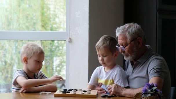 Crianças jogam damas com o avô em casa, homem positivo sênior — Vídeo de Stock