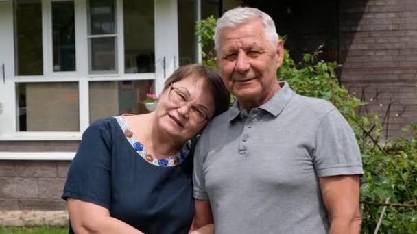 Feliz pareja de ancianos - anciano hombre y mujer mirando a la cámara, al aire libre, casa en el fondo — Vídeos de Stock