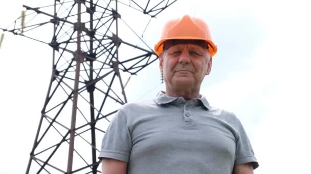 Engenheiro-chefe do homem sênior no chapéu duro olhando para a câmera, pela torre de transmissão, linhas elétricas — Vídeo de Stock