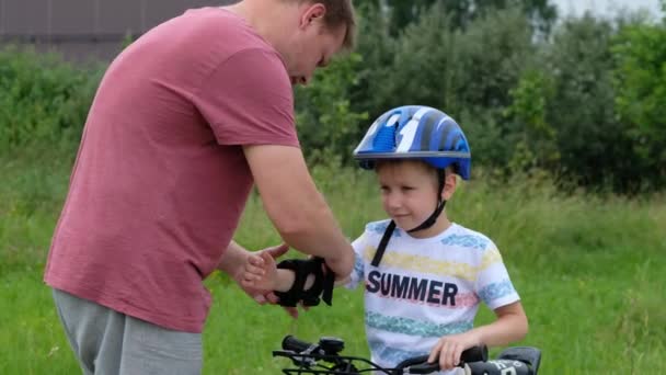 Omtänksam far sätter armbåge pad på sina små söner huvud sedan lära glad pojke att cykla — Stockvideo