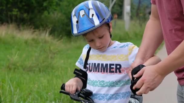 Omtänksam far sätter armbåge pad på sina små söner huvud sedan lära glad pojke att cykla — Stockvideo