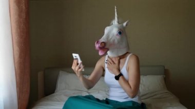 Kafasında tek boynuzlu at maskesi olan bir kadın yatak odasındaki akıllı telefondan web kamerasıyla konuşuyor. Sosyal medya konsepti. Tuhaf komik video.