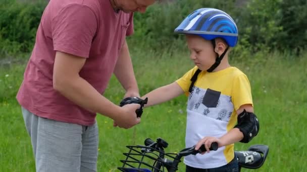 Caring father is putting elbow pad on his little sons head then teaching happy boy to ride bicycle — Stock Video