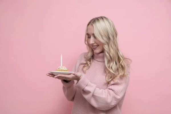 Happy blond woman in pink sweater holding cake with candles, celebrating birthday on pastel pink background. Copy space