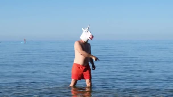 Homem de shorts vermelhos com unicórnio cabeça máscara dançando no mar, estranho engraçado estranho vídeo. — Vídeo de Stock