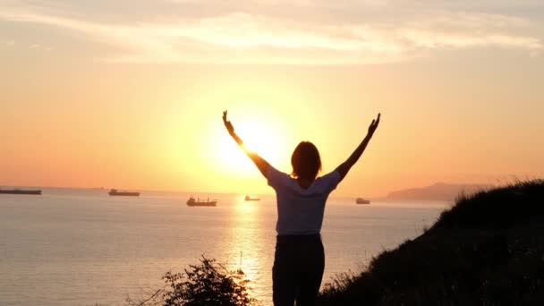 Silhouette d'une femme écartant les bras pour donner à l'océan calme et à l'horizon orange une accolade chaleureuse, plan central, vue de dos. — Video