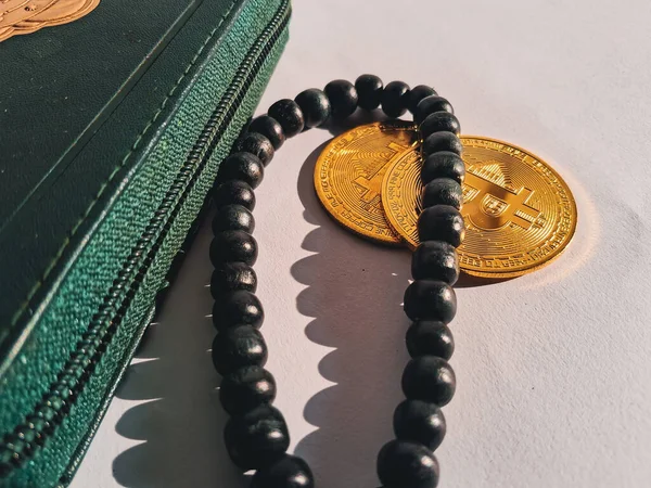 golden Bitcoin coins placed beside a green wallet and black prayer beads (tasbih) on white background