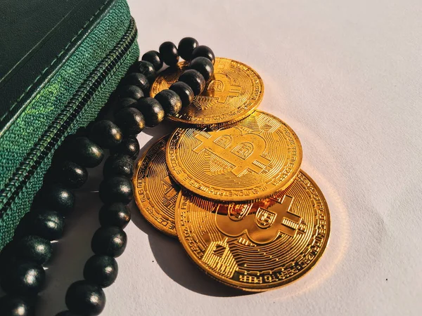 golden Bitcoin coins placed beside a green wallet and black prayer beads (tasbih) on white background