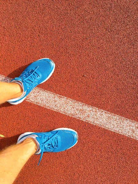 Stadyumun kırmızı koşu bandında mavi spor ayakkabılı bir adam koşuyor.