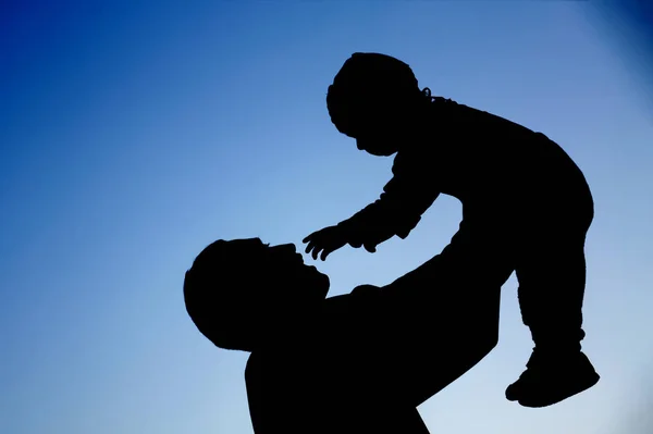 Silhouettes of father and baby son which are playing against the blue sky background. Concept of summer vacation, loving and friendly family. — Stock Photo, Image