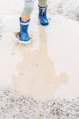 Mavi çizmeli çocuk ve pastel elbiseli çocuk çamurlu su birikintisine basıyor çakıl yolda, doğal ışık, açık hava oyunu ve yağmurlu gün eğlencesi konsepti