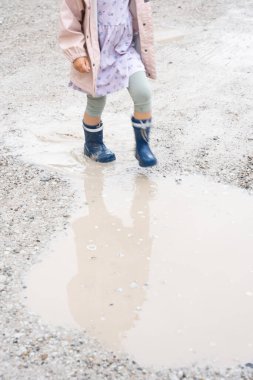 Mavi çizmeli çocuk ve pastel elbiseli çocuk çamurlu su birikintisine basıyor çakıl yolda, doğal ışık, açık hava oyunu ve yağmurlu gün eğlencesi konsepti