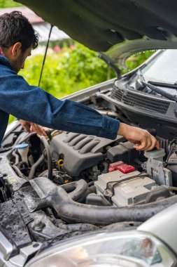 Adam açık kaputun altında motor yağını inceliyor, gerçekçi fotoğraf stili, yağmurlu bir günde açık havada park edilmiş araba, araç bakımı kavramı.