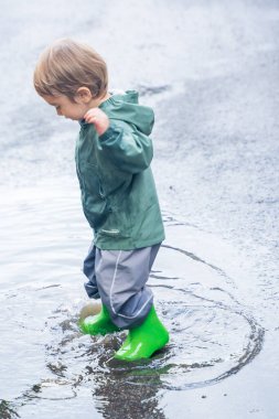 Yeşil yağmurluklu ve botlu bir çocuk ıslak asfalt bir yolda çömelmiş ve su birikintisinde oynuyor, açık havada doğal ışık altında yakalanmış.