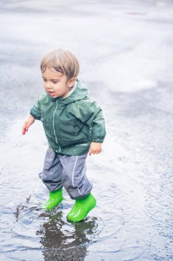 Su birikintisinde sıçrayan çocuk, su geçirmez giysiler ve botlar, doğal açık hava düzeni, eğlence ve çocukluk macerası konsepti.