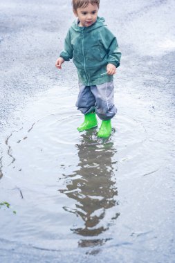 Su birikintisinde sıçrayan çocuk, su geçirmez giysiler ve botlar, doğal açık hava düzeni, eğlence ve çocukluk macerası konsepti.
