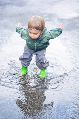 Su birikintisinde sıçrayan çocuk, su geçirmez giysiler ve botlar, doğal açık hava düzeni, eğlence ve çocukluk macerası konsepti.