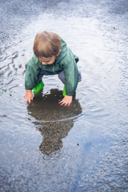 Yeşil yağmurluklu ve botlu bir çocuk ıslak asfalt bir yolda çömelmiş ve su birikintisinde oynuyor, açık havada doğal ışık altında yakalanmış.