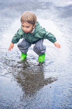 Su birikintisinde sıçrayan çocuk, su geçirmez giysiler ve botlar, doğal açık hava düzeni, eğlence ve çocukluk macerası konsepti.