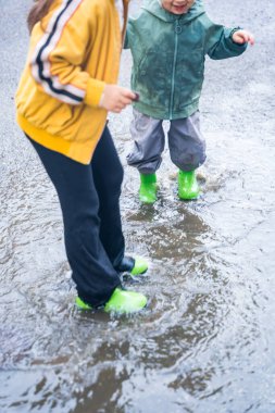 İki çocuk yağmur birikintisinde, yeşil çizmeli renkli giysiler içinde ıslak bir şehir sokağında, açık hava eğlencesi konseptinde zıplıyorlar.