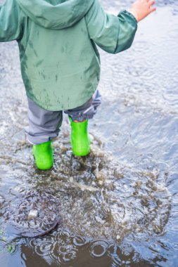 Yeşil çizme giyen ve su geçirmez pantolon giyen bir çocuk su birikintisinde duruyor, doğal tarzda, ıslak asfaltta, eğlenceli yağmurlu gün konsepti
