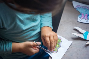 Küçük bir çocuk renkli kağıt bebek kıyafetlerini kesmek için makas kullanıyor, doğal gün ışığı, tahta bir masanın üzerinde. Çocukların yaratıcılık ve el sanatları kavramı