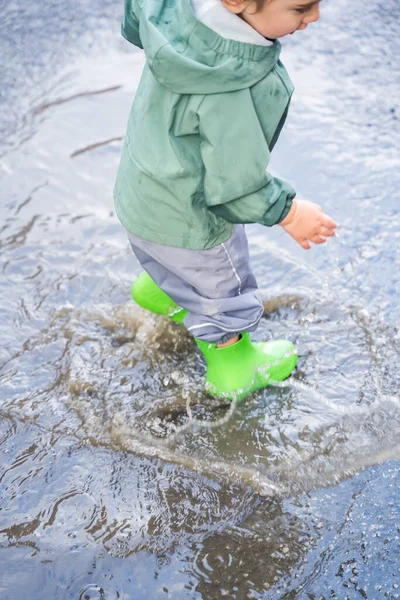 Yeşil çizme giyen ve su geçirmez pantolon giyen bir çocuk su birikintisinde duruyor, doğal tarzda, ıslak asfaltta, eğlenceli yağmurlu gün konsepti