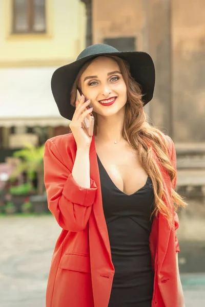 かわいいファッショナブルな若い女性が帽子と赤いジャケットを着て路上で電話で話しています — ストック写真