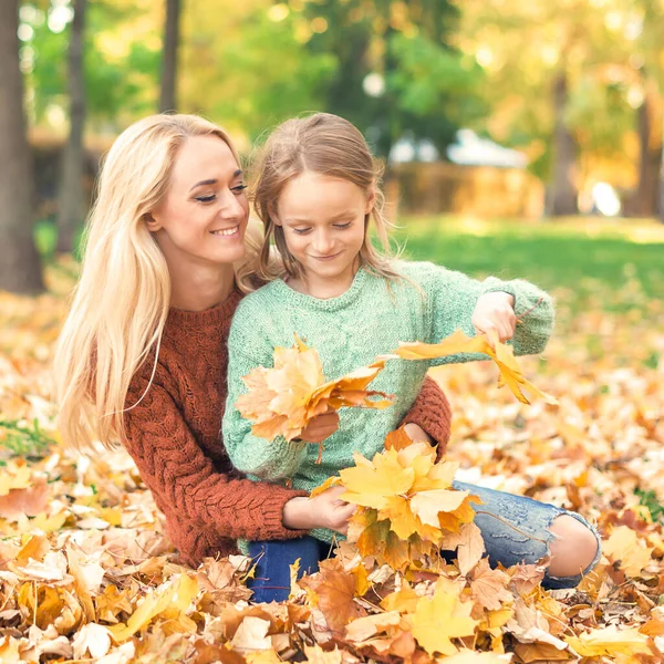 快乐的年轻的高加索母亲和小女儿抱着秋天的黄叶坐在公园里 — 图库照片