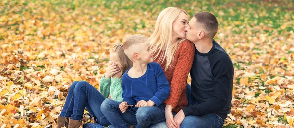 年轻家庭坐在秋天树叶上的画像 在秋天的公园里 父母亲吻和与孩子们坐在一起 — 图库照片