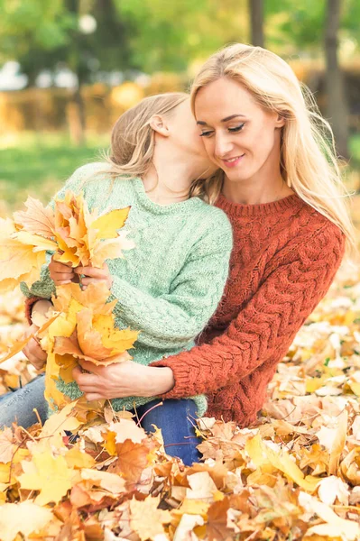 快乐的年轻的高加索母亲和小女儿抱着秋天的黄叶坐在公园里亲吻 — 图库照片