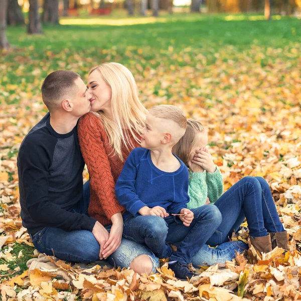 年轻家庭坐在秋天树叶上的画像 在秋天的公园里 父母亲吻和与孩子们坐在一起 — 图库照片