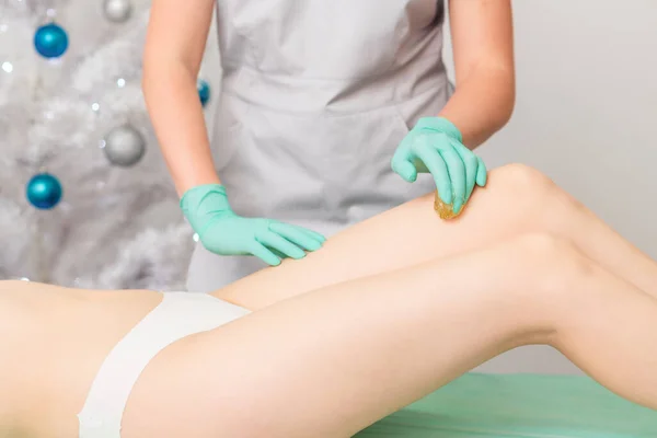 Beautician waxing legs of woman in spa center.