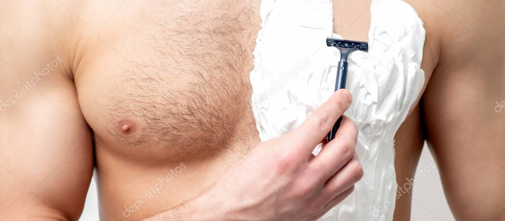 Young white man holds razor shaves his chest with white shaving foam on white background. Man shaving his torso