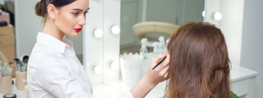 Makyaj sanatçısı müşterisiyle güzellik salonundaki aynanın yanında çalışıyor.