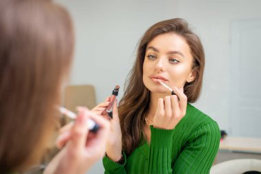 Güzel beyaz kadın aynaya bakarak dudaklarına parlatıcı sürüyor.