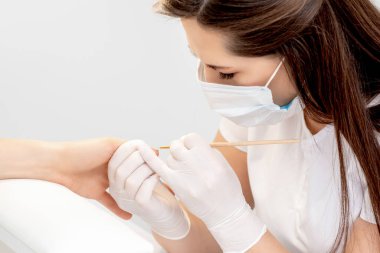 Plastik eldivenli koruyucu maskeli manikür ustası güzellik salonundaki kadın tırnaklarına bej oje sürüyor.