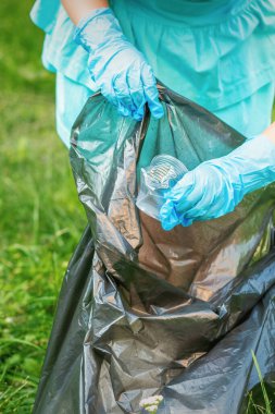 Çocuk çimenlerden plastik çöp topluyor. Parktaki çöp torbasına çöp atıyor.