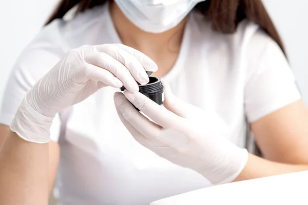 Beautician or manicure master with protective mask holds black jar with cosmetics or manicures product in her hands with white gloves