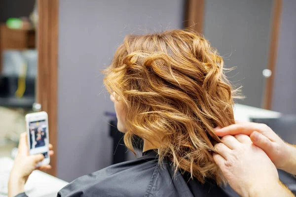 Fryzjer Robi Fryzurę Dla Młodej Kobiety Czerwonymi Kręconymi Włosami Smartfonem — Zdjęcie stockowe