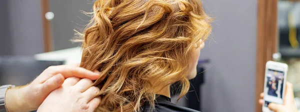 Fryzjer Robi Fryzurę Dla Młodej Kobiety Czerwonymi Kręconymi Włosami Smartfonem — Zdjęcie stockowe