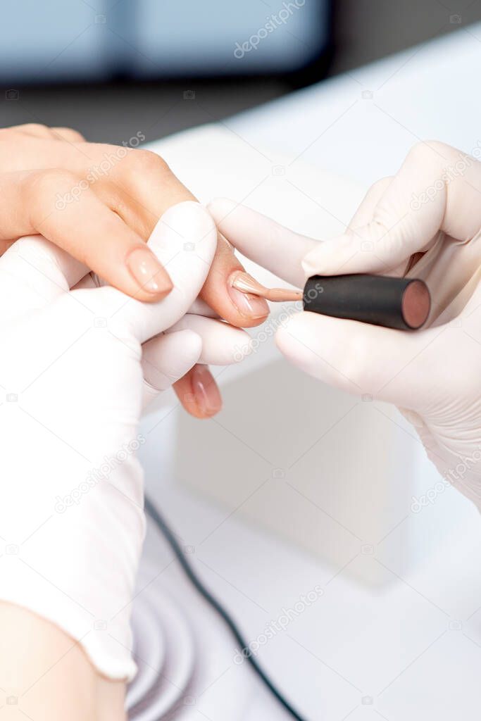 Manicure master in protective gloves applying beige nail polish on female nails in beauty salon