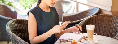 Genç iş kadını, akıllı telefondan not defterine kahve molasında çalışırken ona bakıyor.