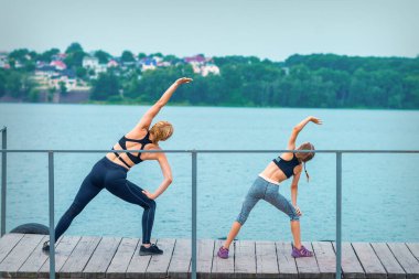 Anne ve kızı nehrin iskelede çimlerin üzerinde egzersiz yapıyor.