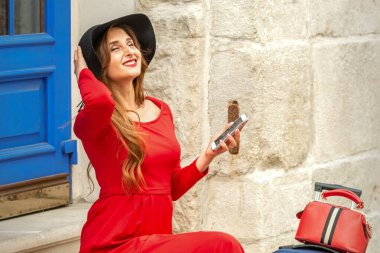 Güzel, genç, beyaz bir kadın kapının yanında oturuyor. Siyah şapkalı, uzun kırmızı elbiseli, seyahat çantası ve akıllı telefonlu.