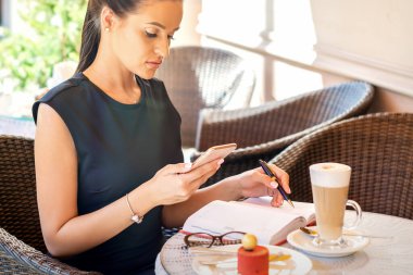 Genç iş kadını, akıllı telefondan not defterine kahve molasında çalışırken ona bakıyor.